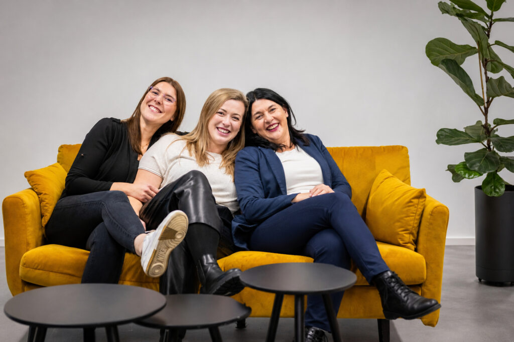 Das Team von Modul Training lacht gemeinsam auf einem gelben Sofa im Büro in Tamm