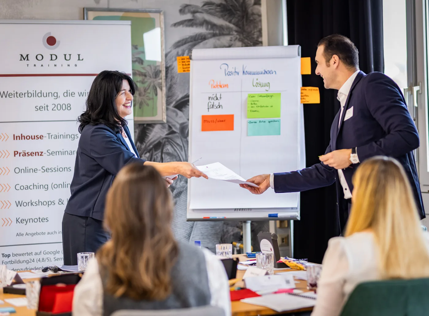 Seminarleitung übergibt im Kommunikationsseminar ein Arbeitsblatt, am Flipchart steht „Positiv kommunizieren“ – MODUL TRAINING
