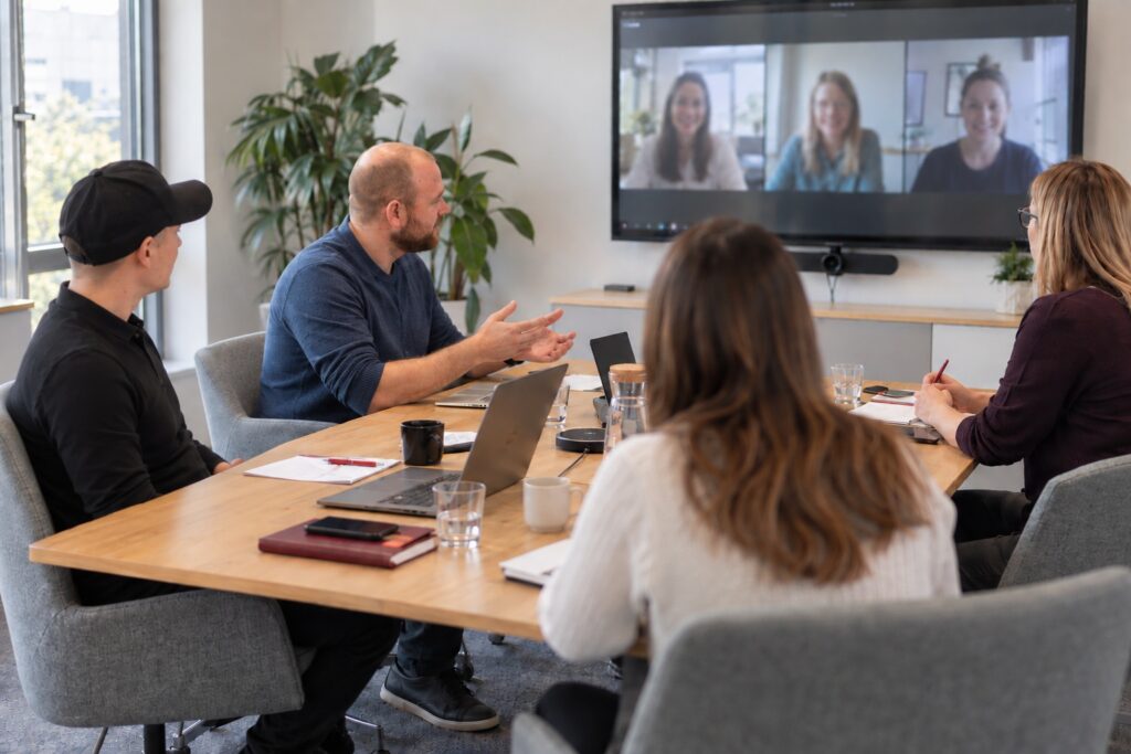 Team im Besprechungsraum, zugeschaltete Kolleginnen auf dem Bildschirm – MODUL TRAINING